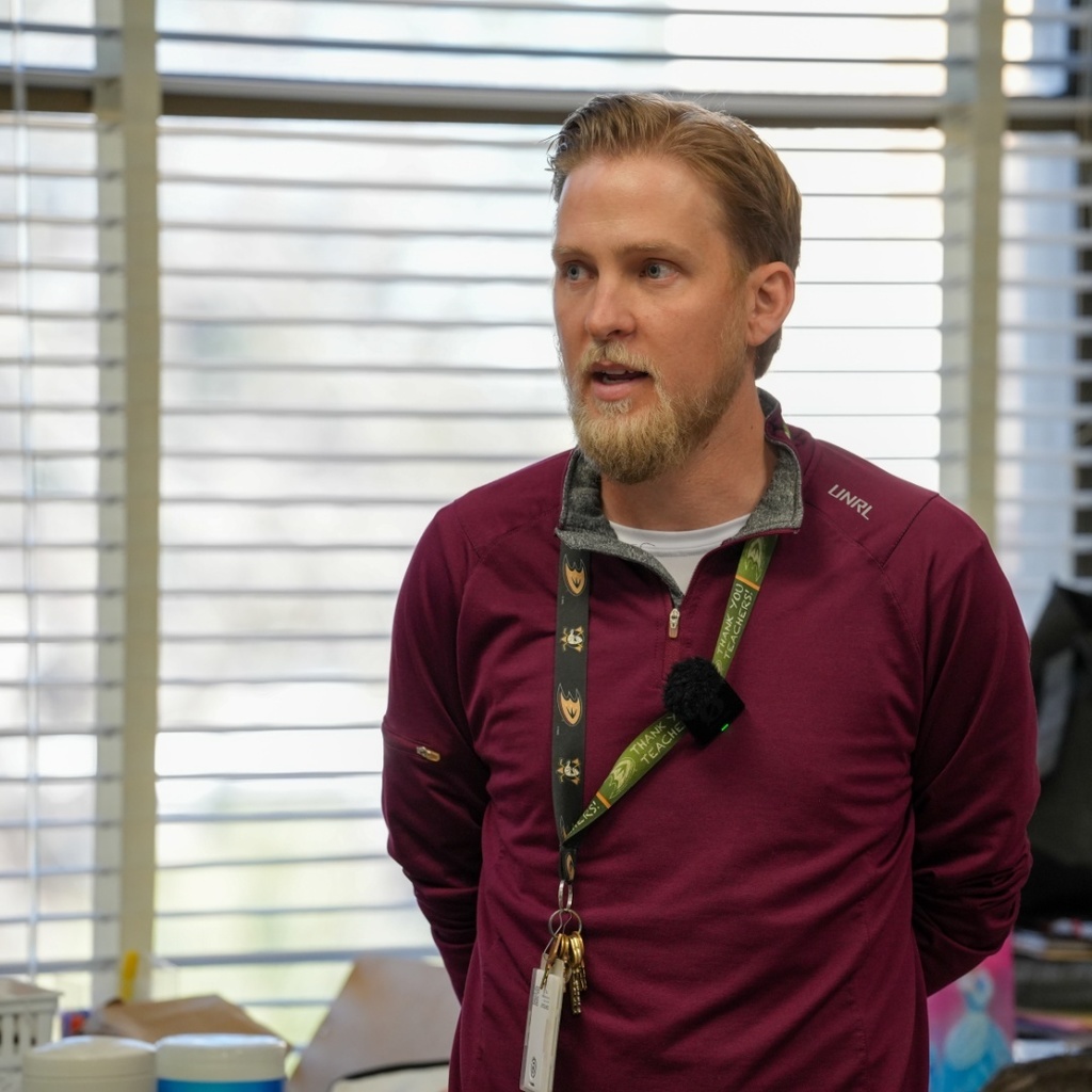 Andrew Ohmannwears a maroon jacket and a lanyard with a microphone. He stands before a desk.