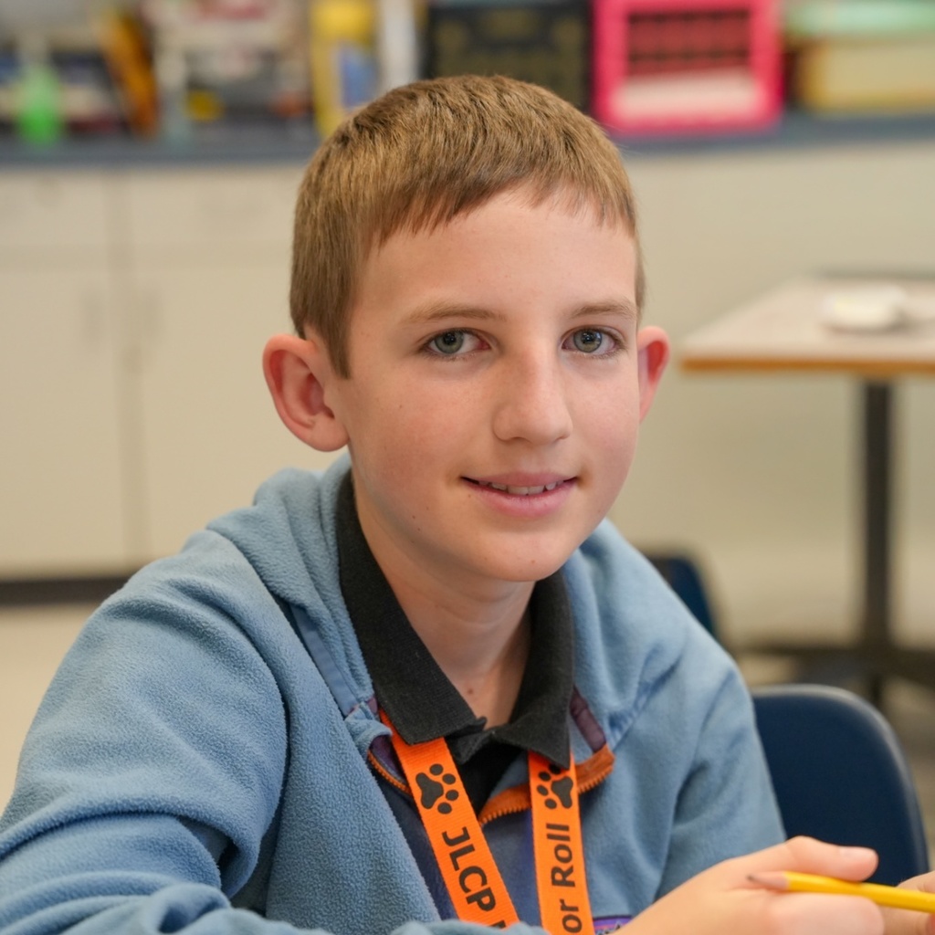 A boy sits in a classroom, holding a yellow pencil. He wears a blue sweater and has an orange lanyard.