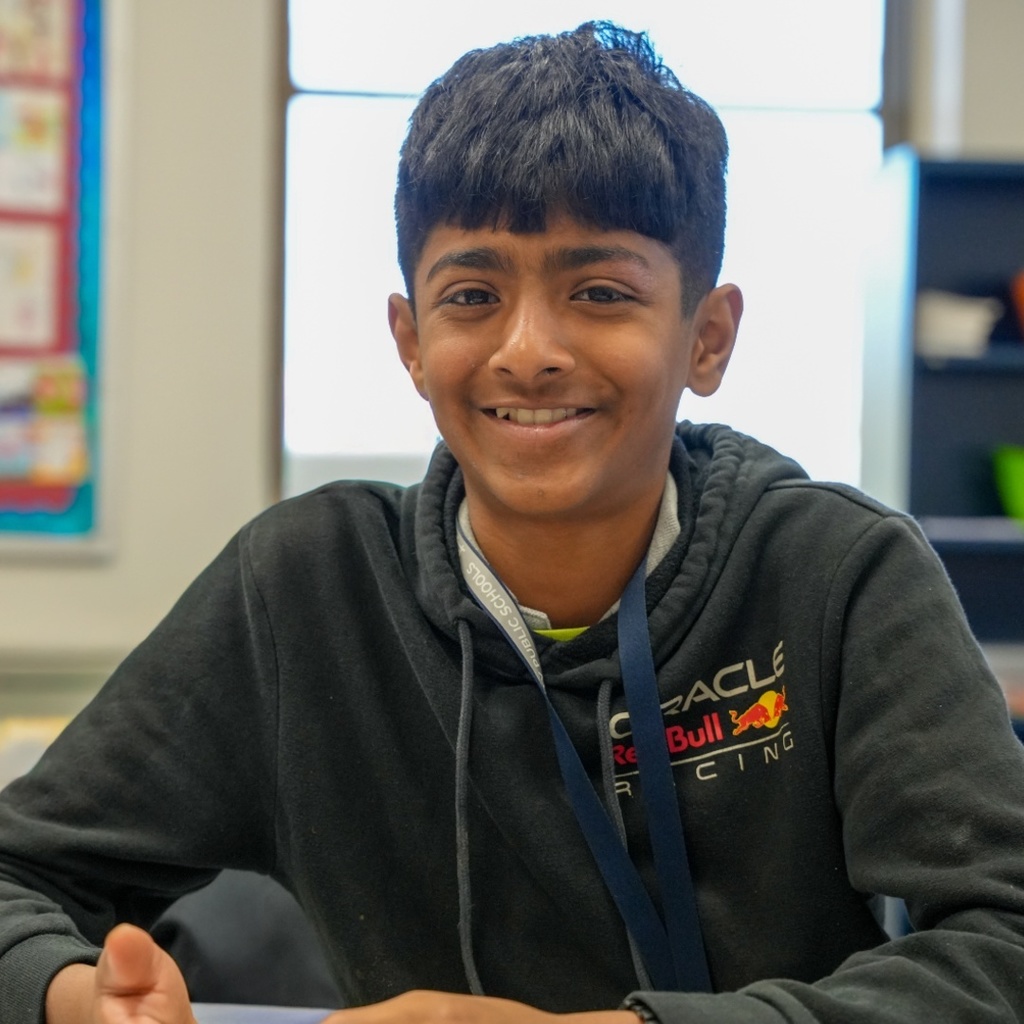 A boy in a black hoodie with "Oracle" on the front sits at a table. He has a smile.