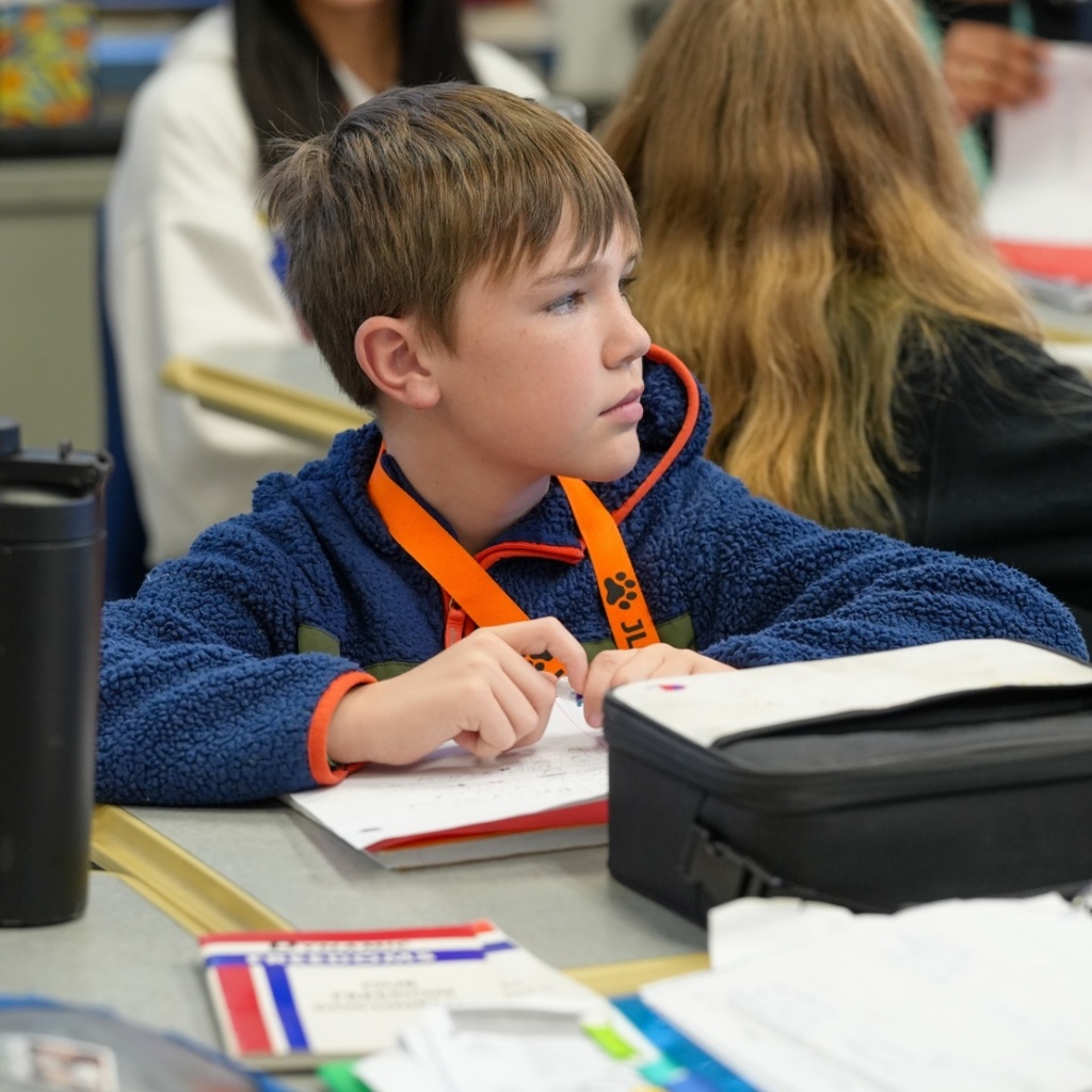 A young boy in a blue hoodie, sitting at a desk, writing in a notebook with other students.