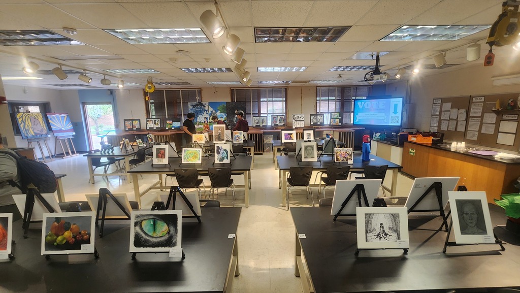 Room with tables, each displaying multiple framed art pieces. A television is mounted on the wall and people are working at the back.