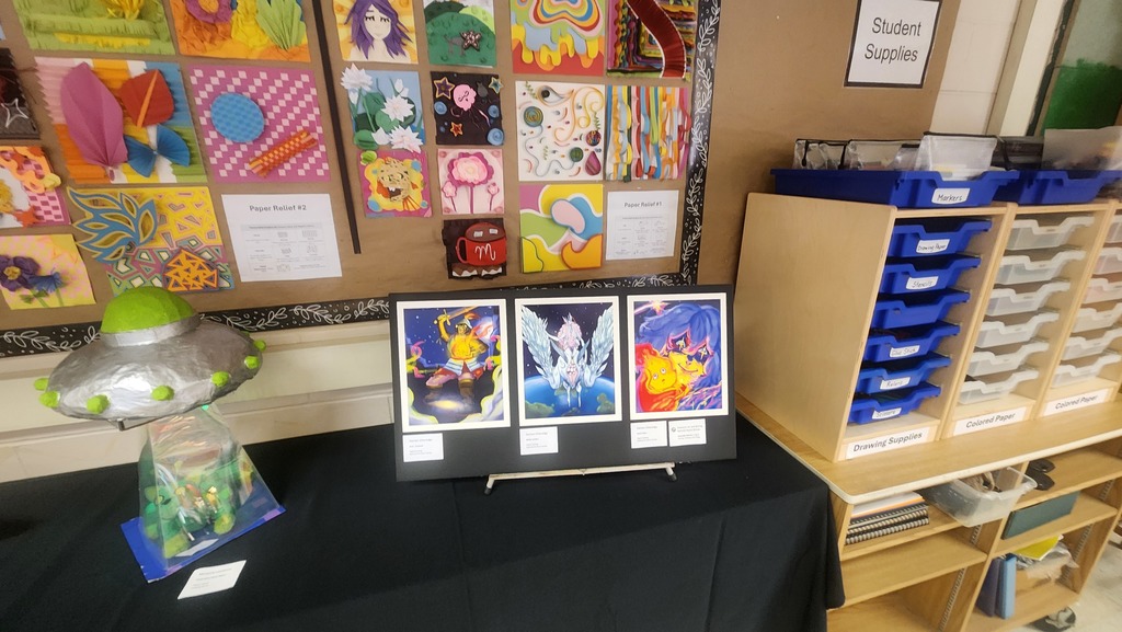 A table with art displays and a clear container with a green lid. A bulletin board is behind the table.