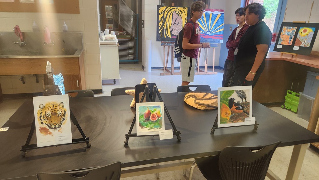 Art display in an art studio, three individuals standing around a table with framed artworks, additional artwork on the wall.