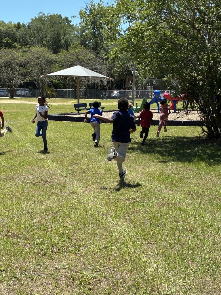 Kids run on a grassy field with trees and a white umbrella in the background.