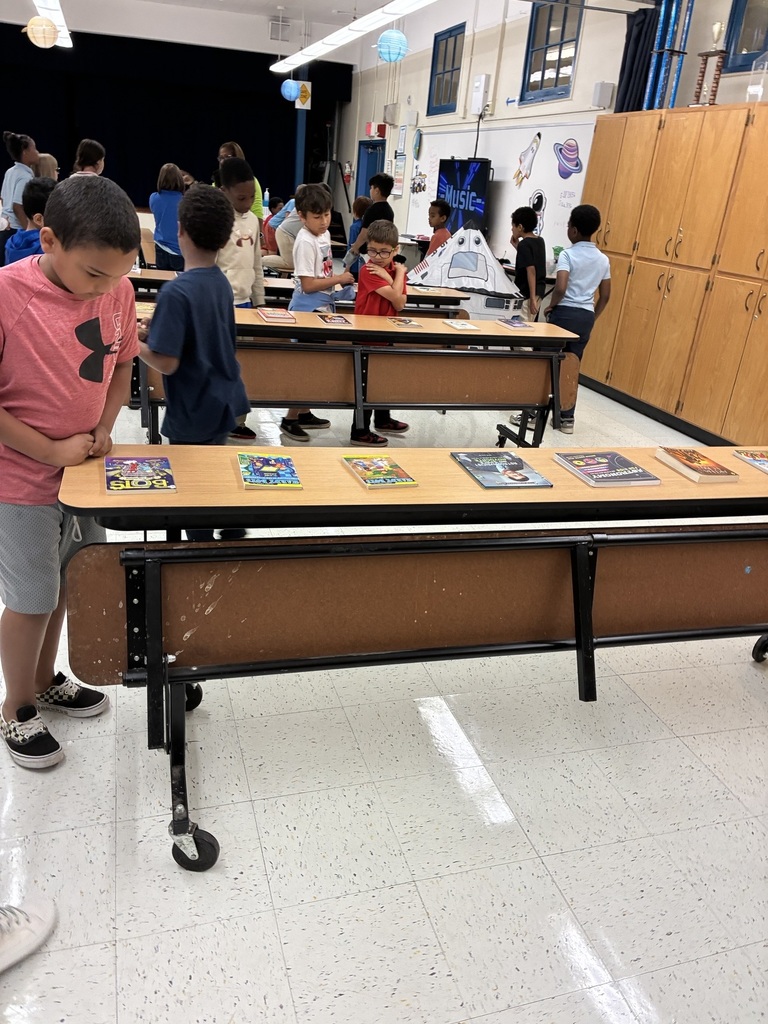 A classroom with desks and children. One child is standing at a desk with books on it.