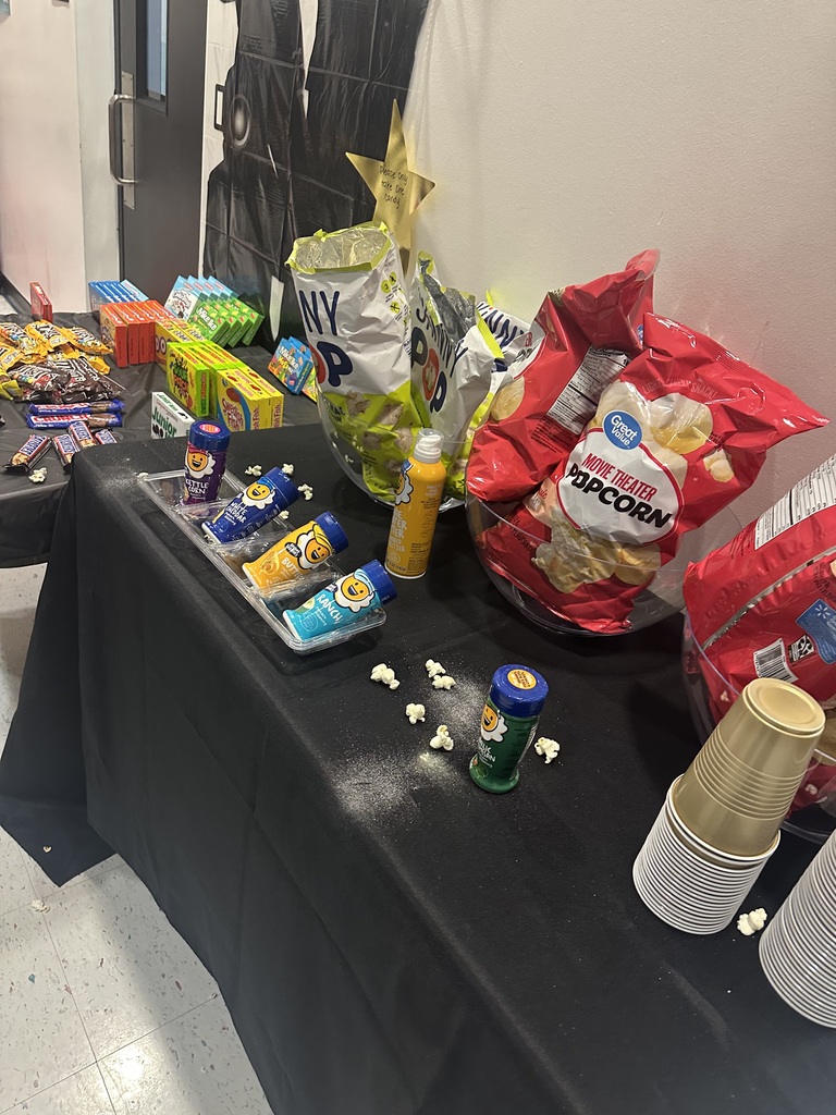 A table with a black tablecloth, covered with snacks like chips, popcorn, drinks, and popcorn kernels.