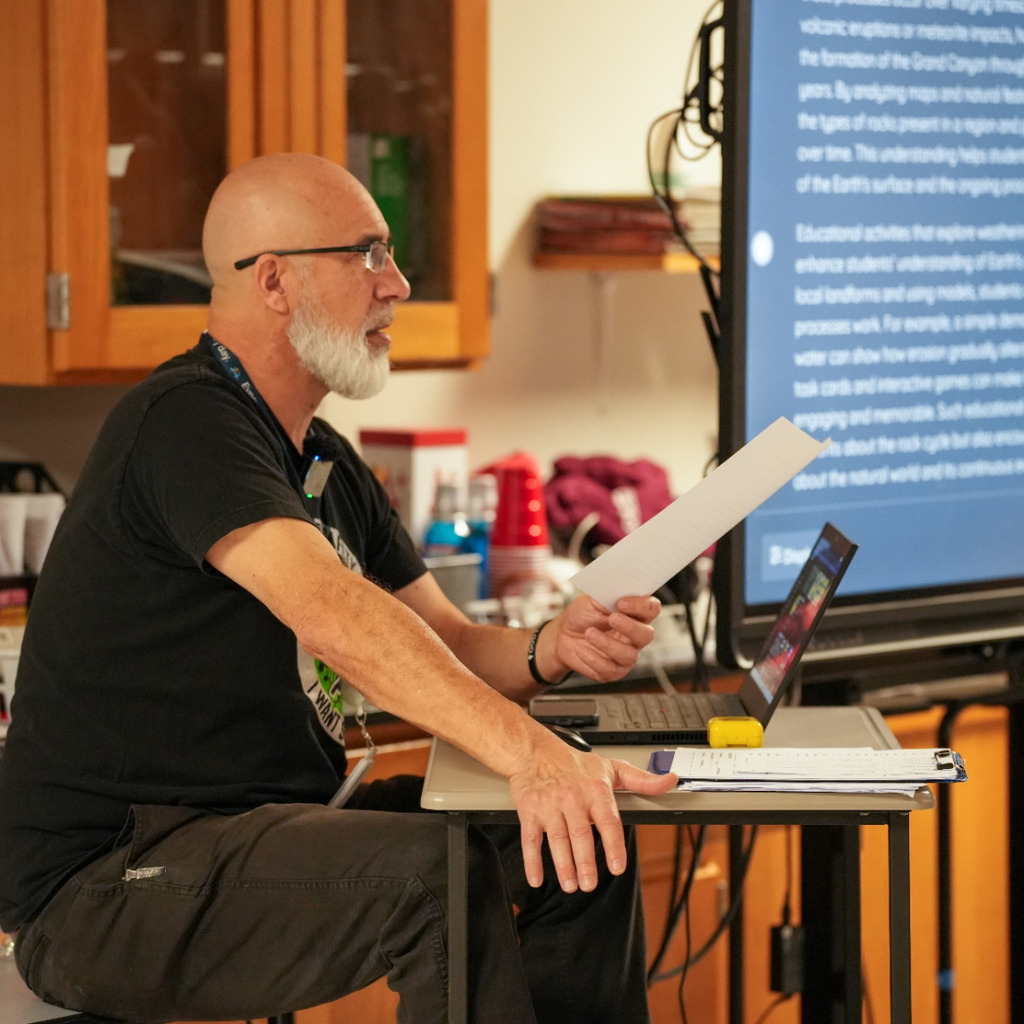 Person in a black shirt seated at a desk, holding a paper, next to a laptop.