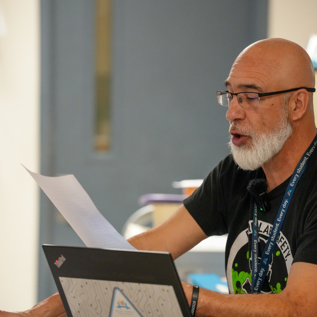 A man with glasses and a lanyard around his neck speaks at a podium with papers in his hand.