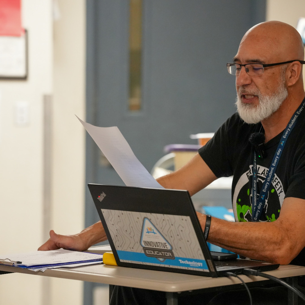 A man sits at a desk with a laptop, papers, and a notebook. He wears glasses and a black shirt.