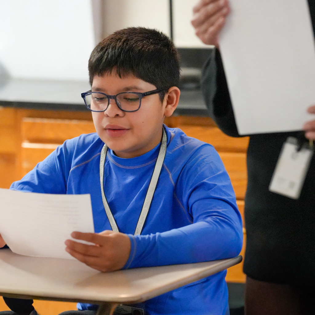A boy with glasses and a blue shirt reads a paper, sitting at a desk in a classroom.