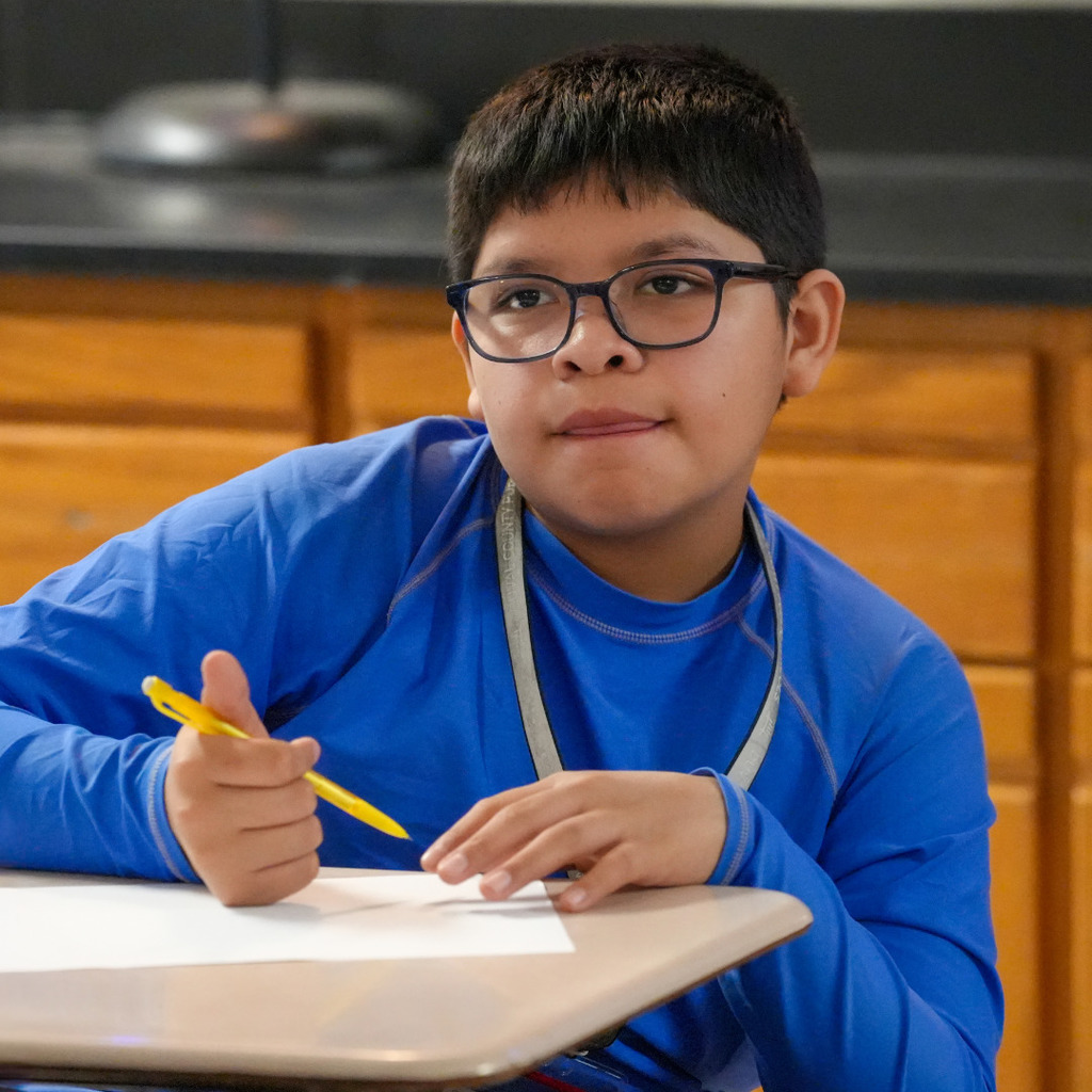 A young boy wearing glasses and a blue shirt sits at a table holding a yellow pencil.