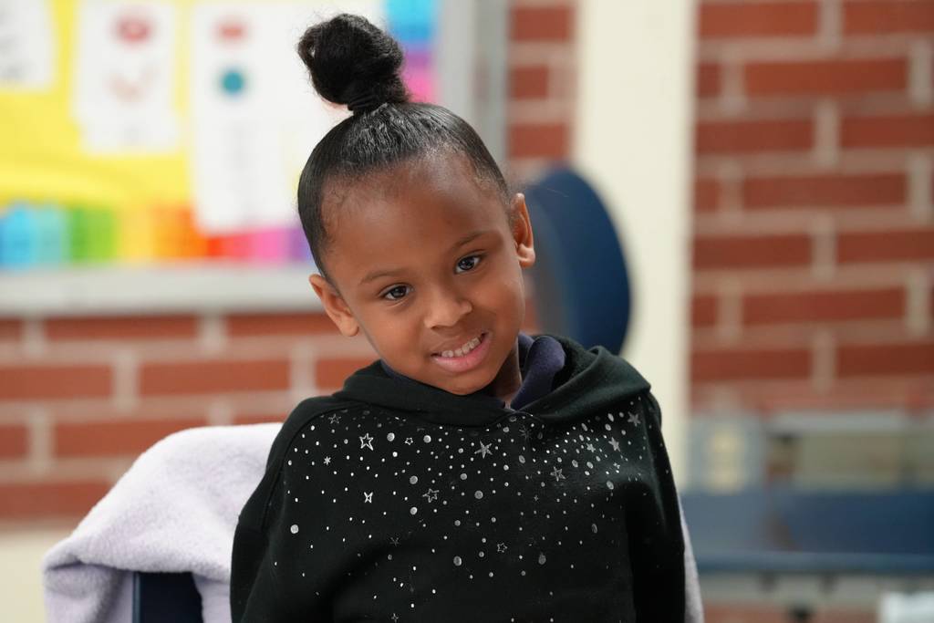 A young girl with a bun hairstyle and a black hoodie with white dots smiles in a classroom.