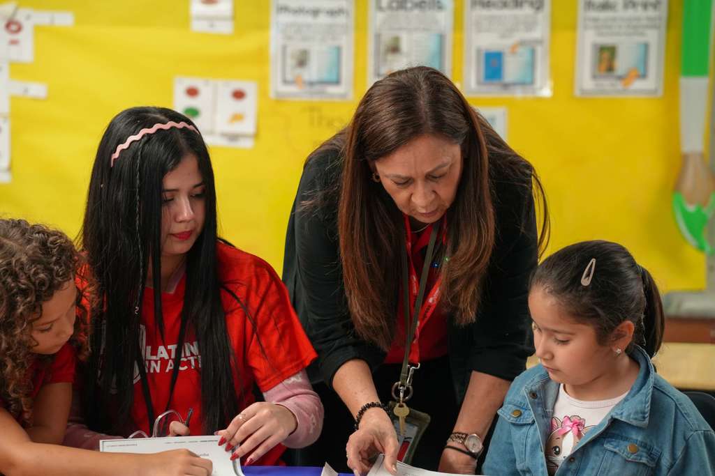 Teacher and students are focused on a book in a classroom with a yellow wall.