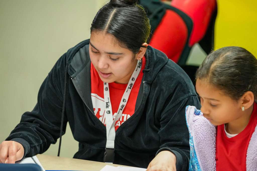 Woman in a black jacket helps a girl in a red shirt with papers on a desk.