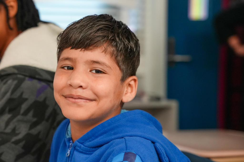 A smiling child in a blue hoodie sits in a classroom, with a person in a camouflage jacket and a blue wall in the background.