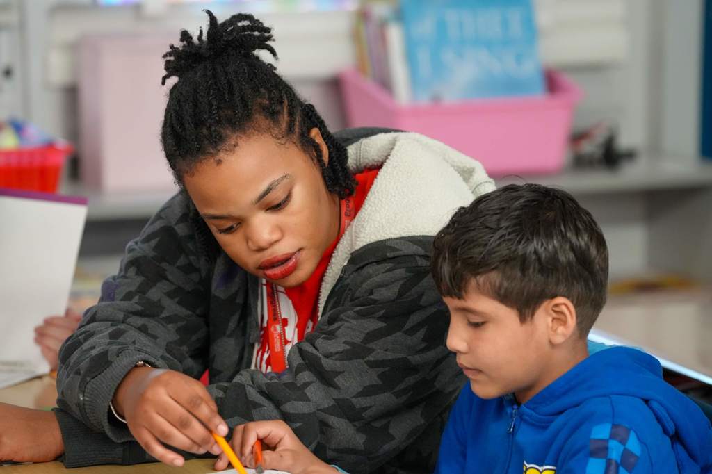 Woman wearing a jacket, helping a child in a blue hoodie with a pencil, sitting at a desk.
