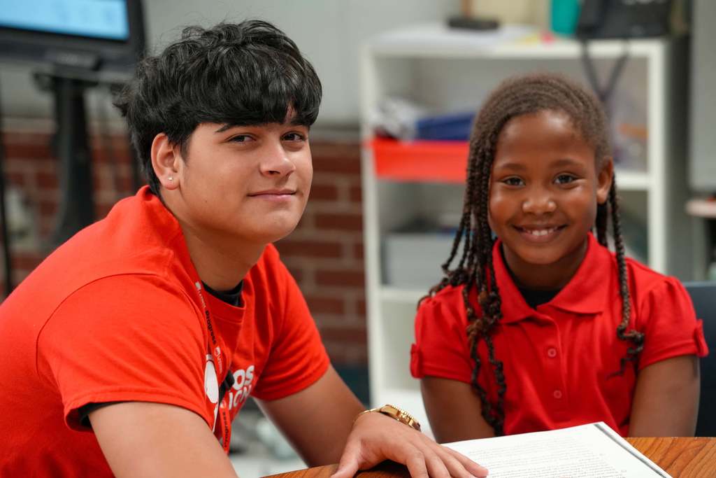 Two students in orange shirts, one seated and smiling, another leaning on a desk with a notebook.