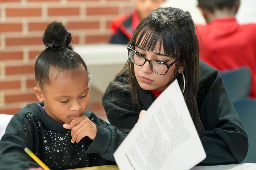 Two students seated, one reading aloud, the other listening, in a classroom with brick walls.
