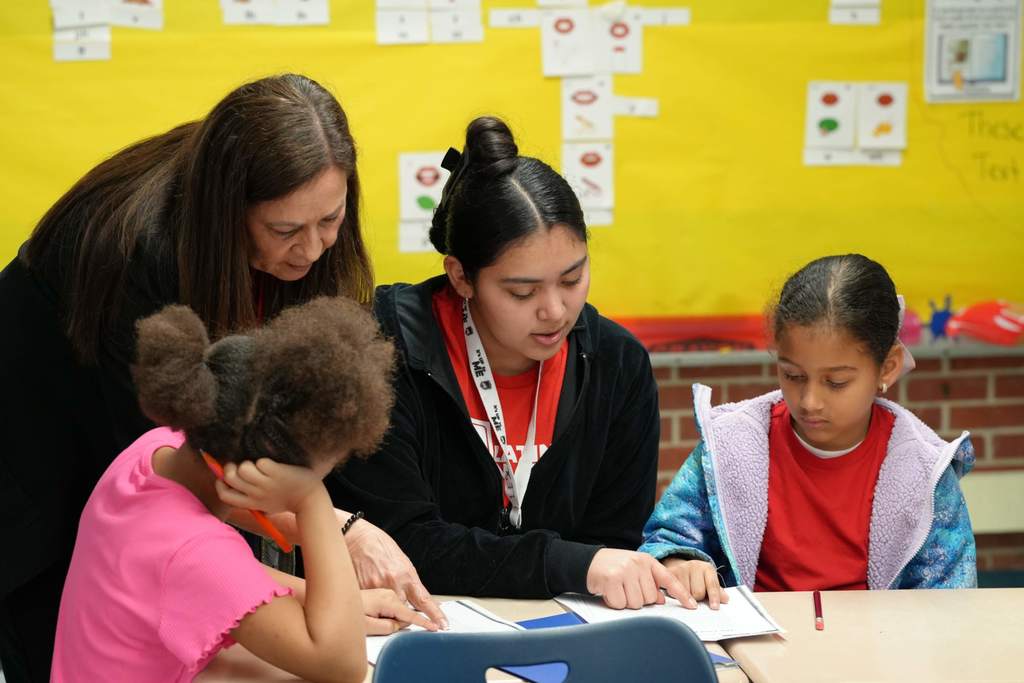 Three children and a woman sit at a table, reviewing papers. The woman wears a lanyard. The room has a yellow wall with colorful shapes.