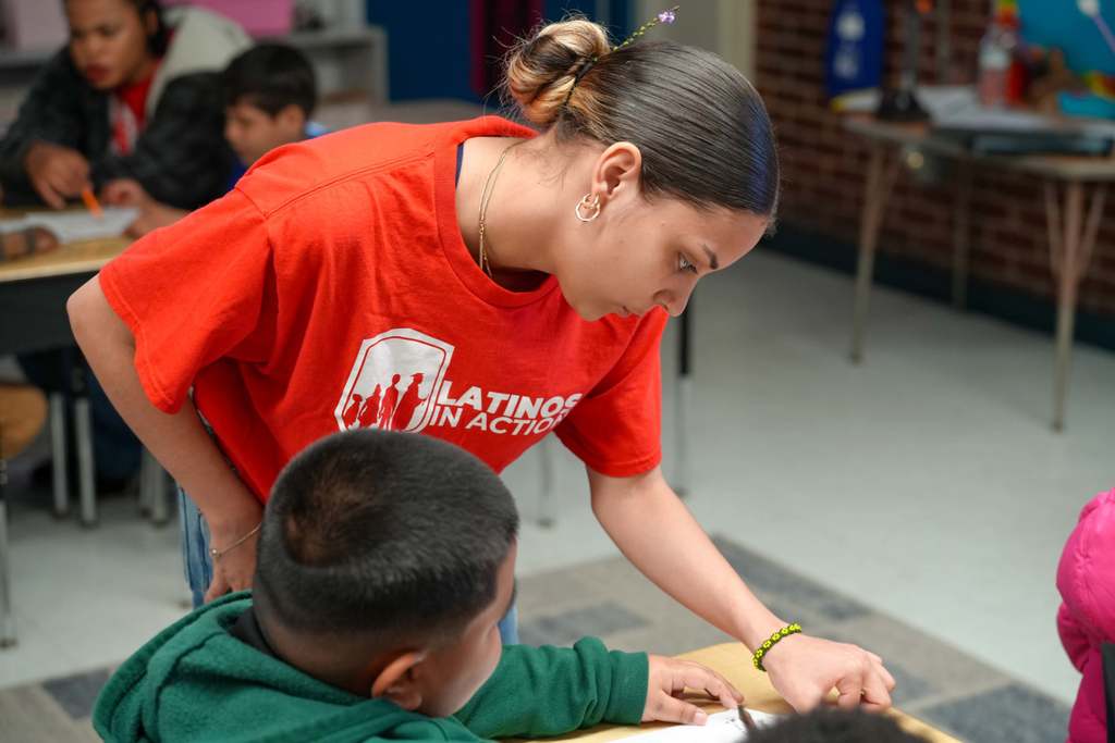 A woman in a red shirt helps a boy in a green shirt at a desk. Behind them, other children work at tables.