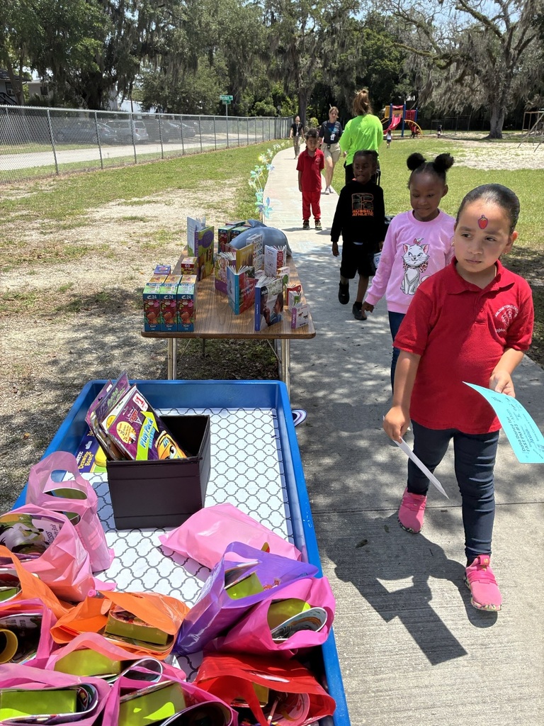 Children walking past a table with books. A child in a red shirt holding a paper.