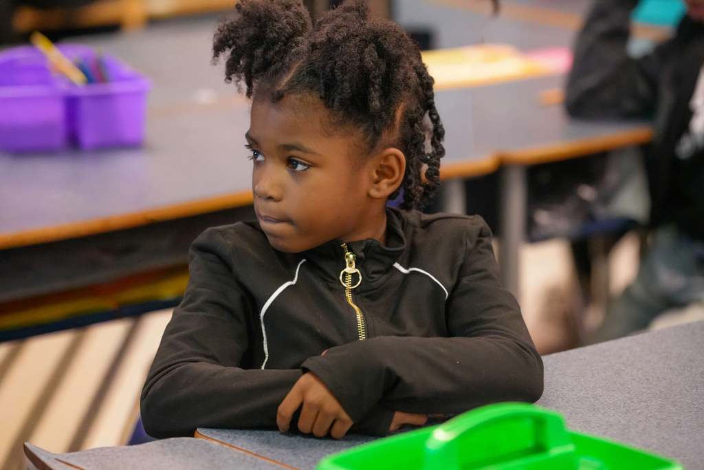 A child sits at a table in a classroom, looking down with folded arms. Purple storage bins and other items are behind them.