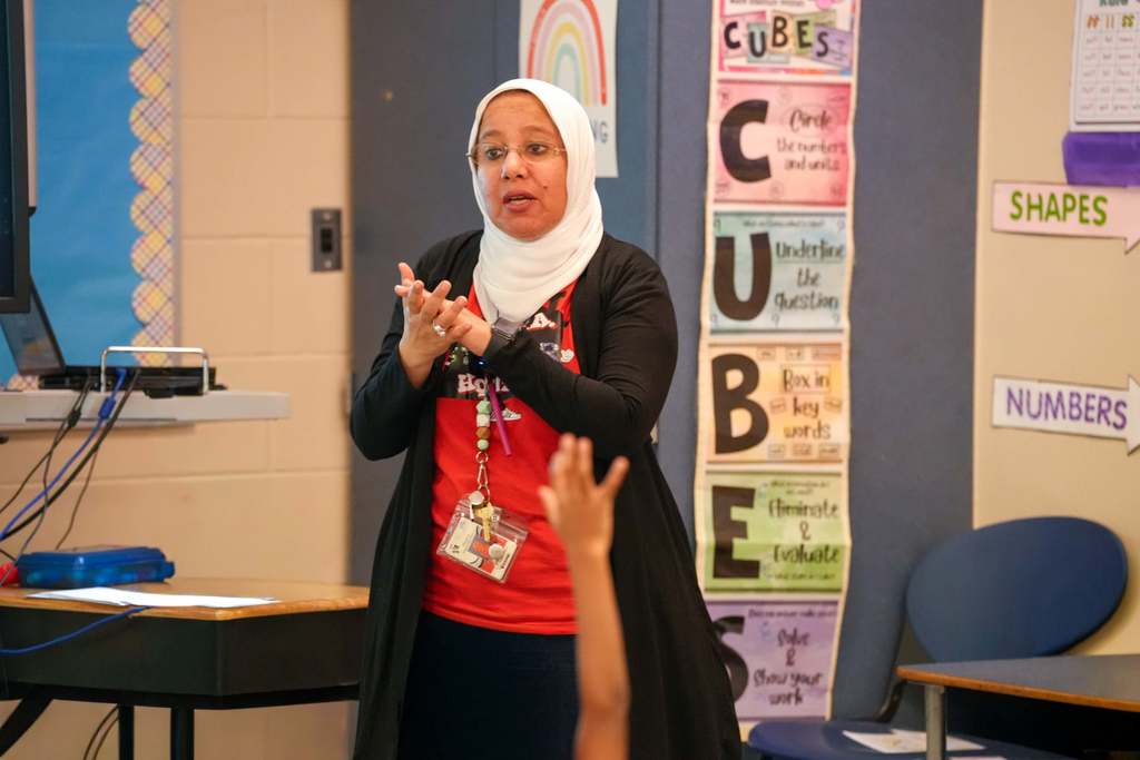 A Ola Reiad teaches in a classroom. A student raises their hand. Colorful posters line the wall.