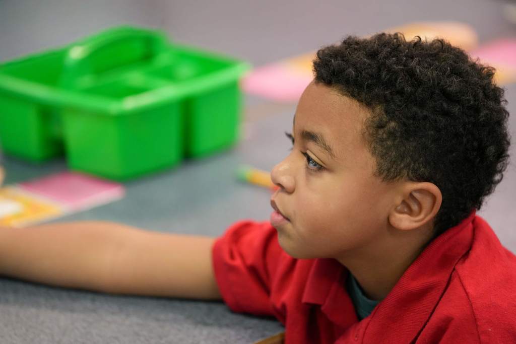 A young boy in a red shirt sits at a table, looking down. Behind him is a green tray with compartments.
