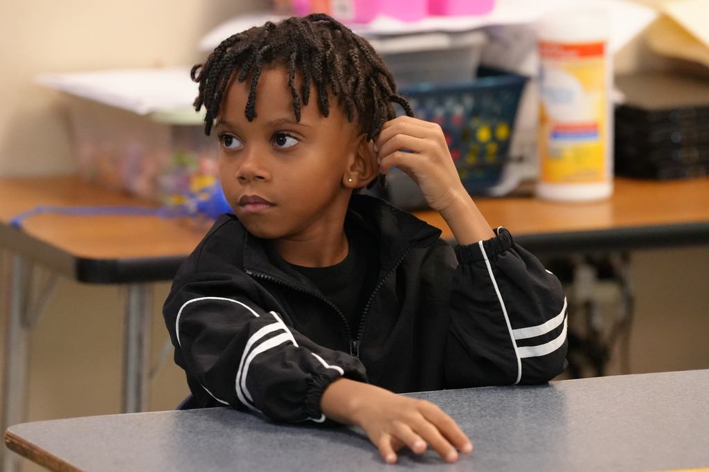 Child seated at desk, wearing a black jacket with white stripes, looking at something with one hand on ear.