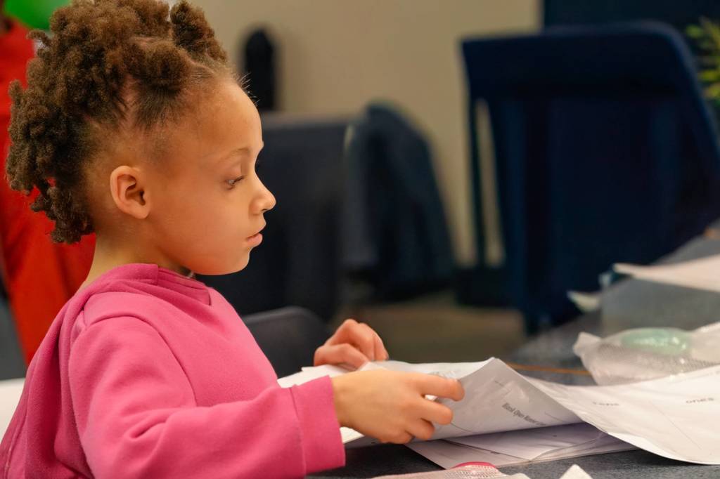 A young child with curly hair, wearing a pink shirt, is sitting and reading a piece of paper.