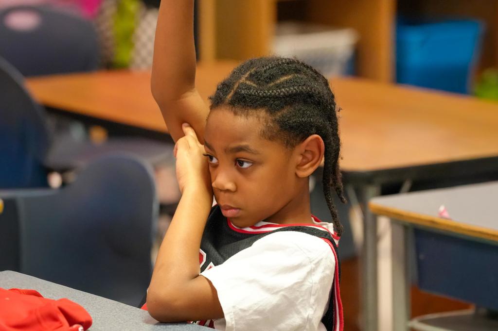A child sits at a table in a classroom, raising an arm and touching her face.