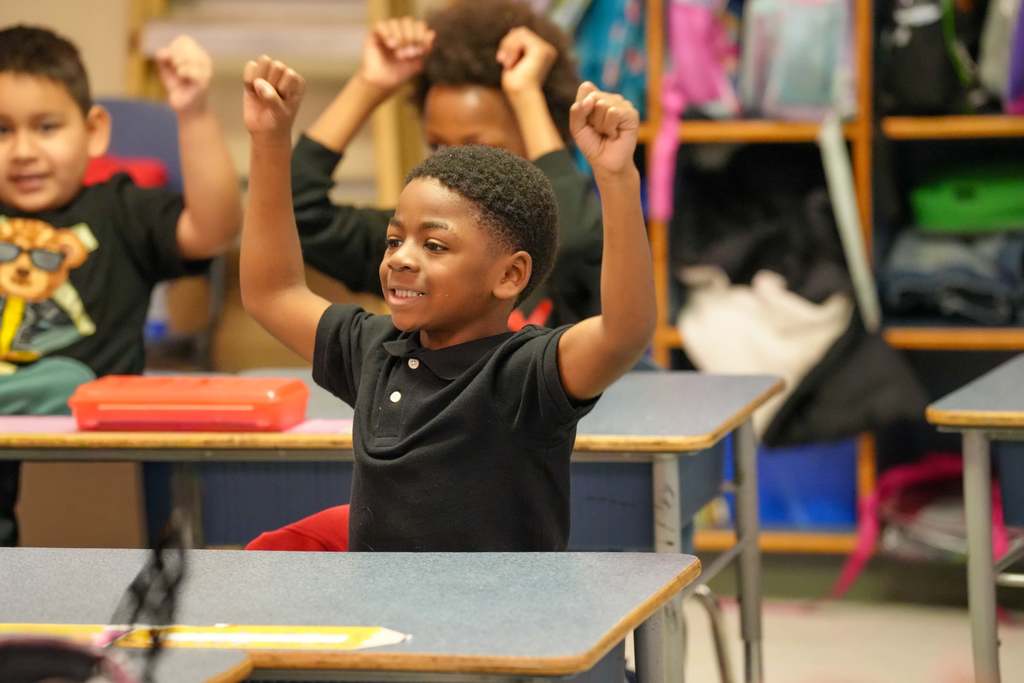Children seated at desks, one boy raises both arms excitedly. Background includes shelves with storage boxes and another student.
