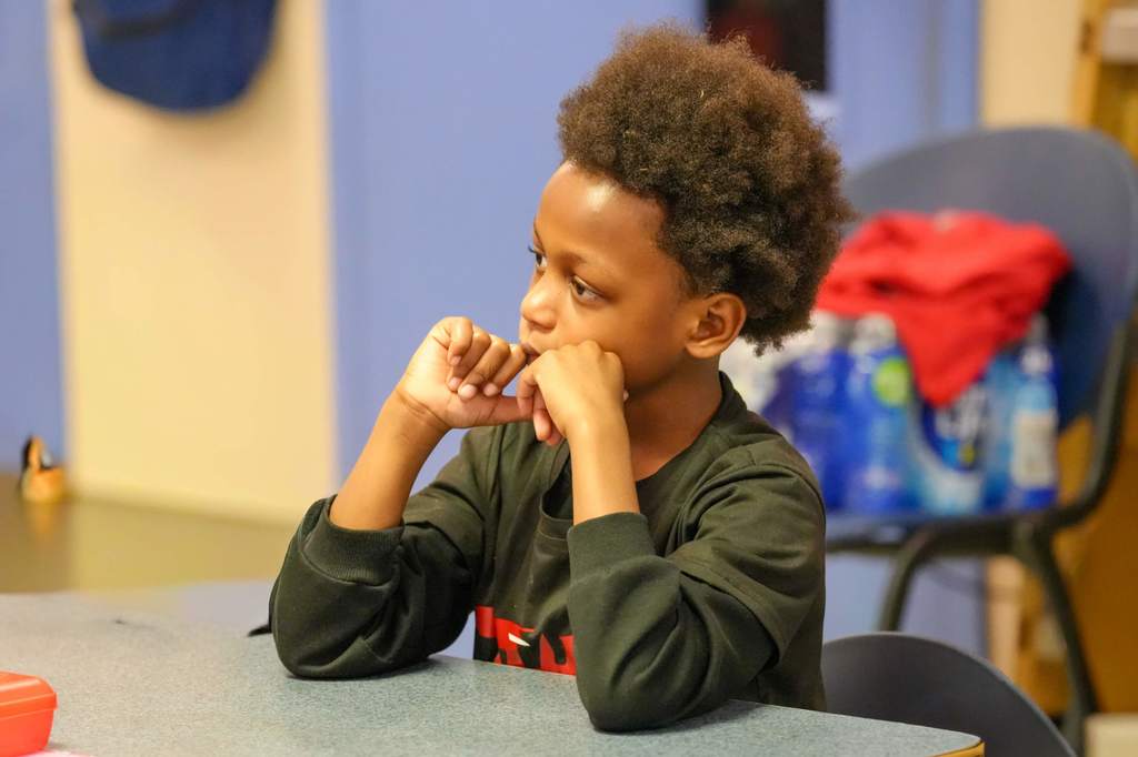A young person sits at a desk, resting their chin on folded hands. They wear a green long-sleeve shirt.