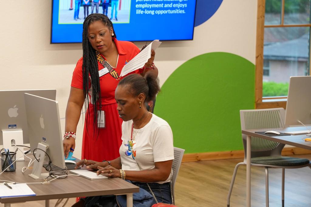 Two women work at a computer in a room with a screen, one standing and one sitting.