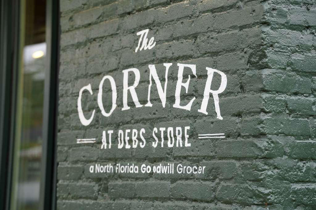 A brick wall with a sign reading "The Corner at Debs Store" in white text, accompanied by reflections of nearby buildings.