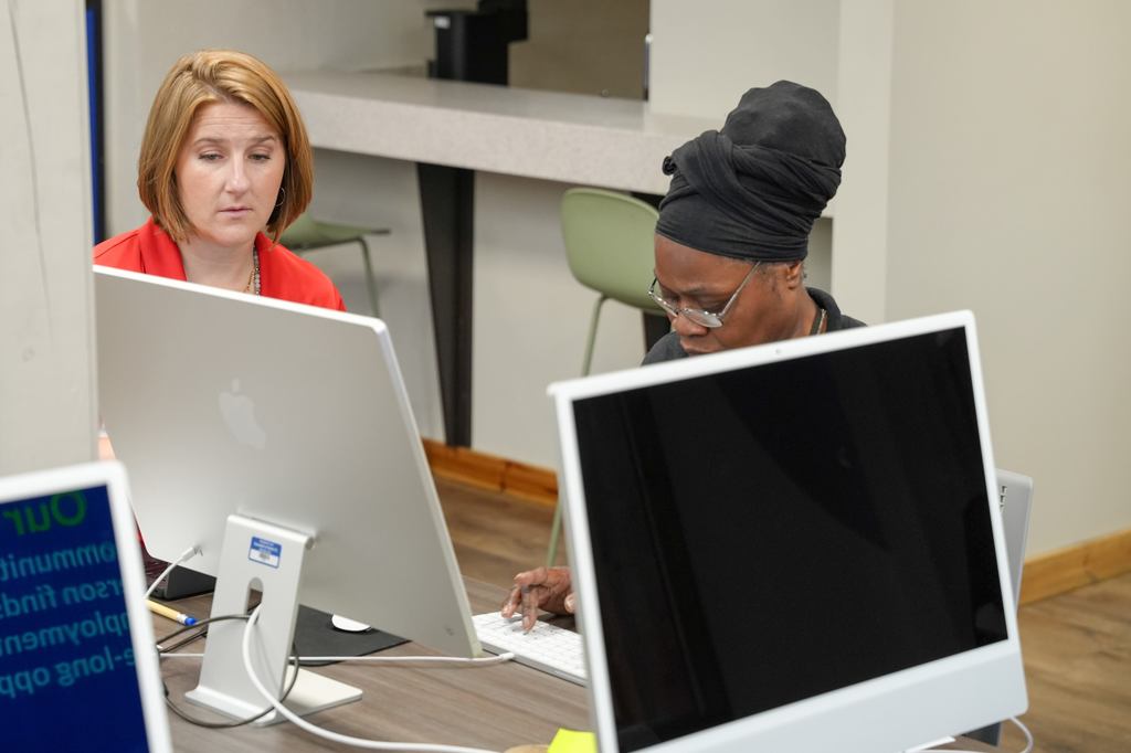 Two women sit at a desk, each using a computer. One woman wears a red top, and the other wears a black hat.