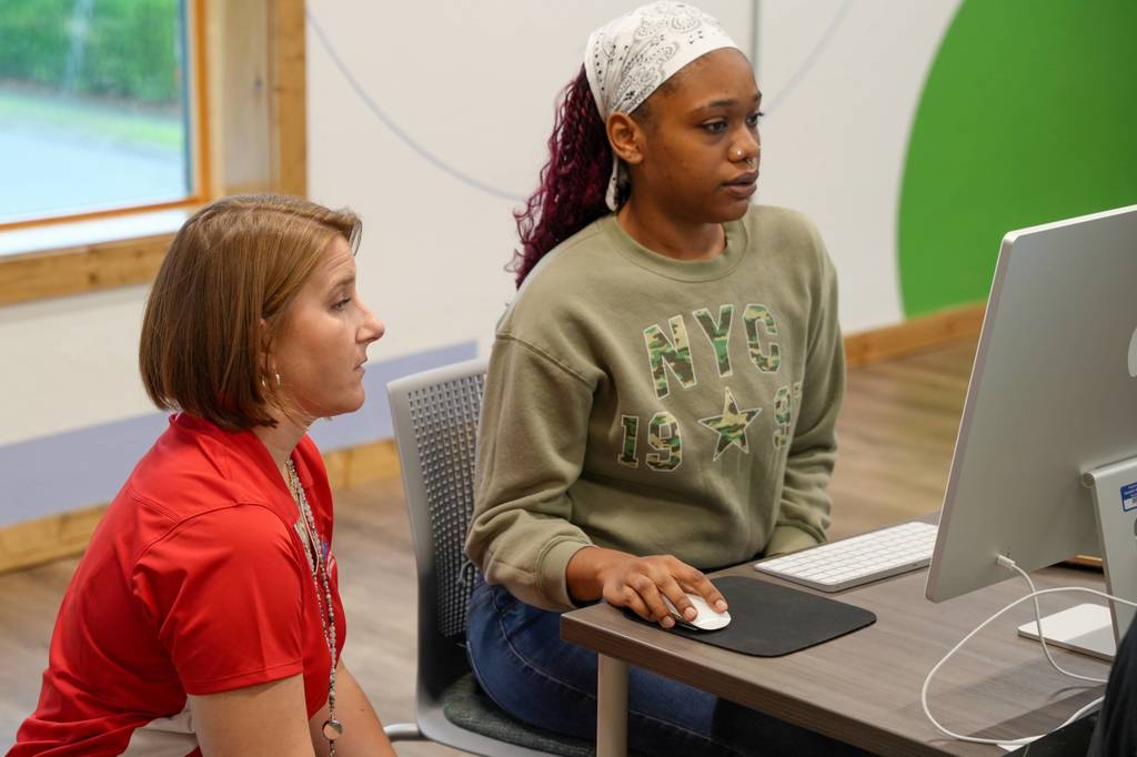 Two women seated at a desk, one using a computer mouse and the other looking at her.