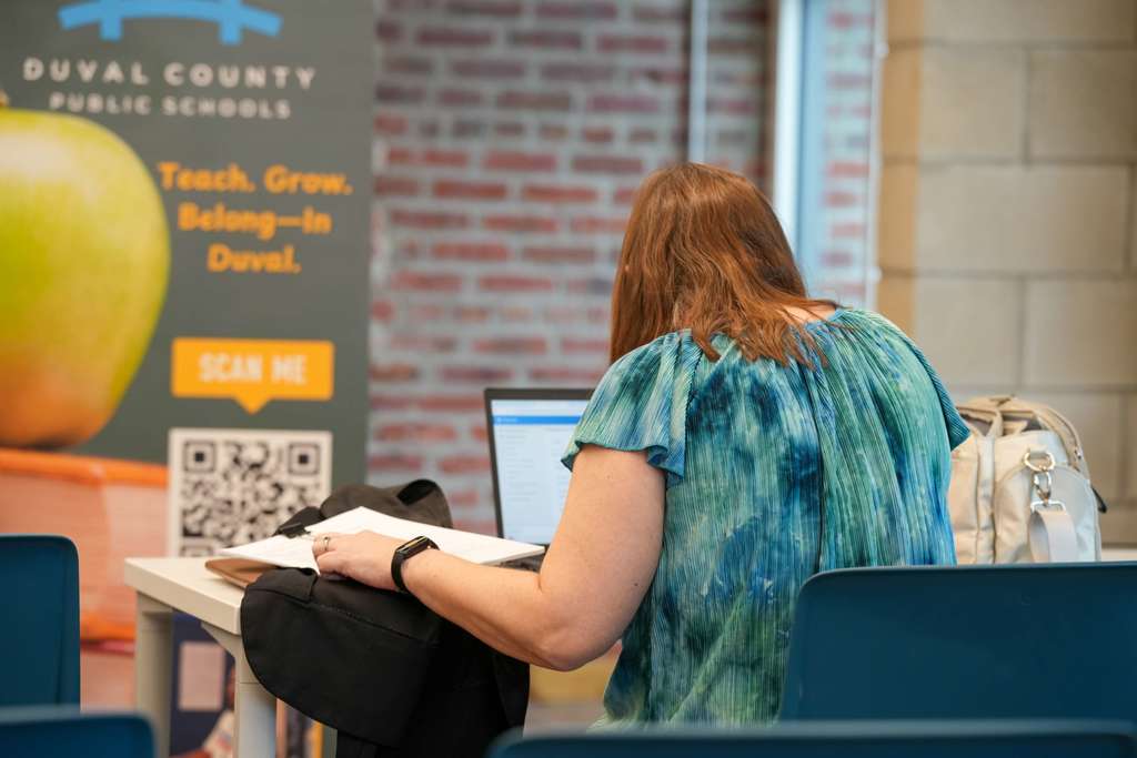 A person in a green top sits at a table with a laptop, near a brick wall and an apple poster.