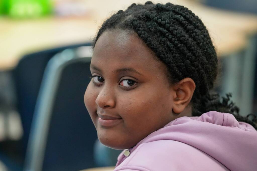 A smiling person with braided hair sits in a chair in a classroom. The background includes a desk.