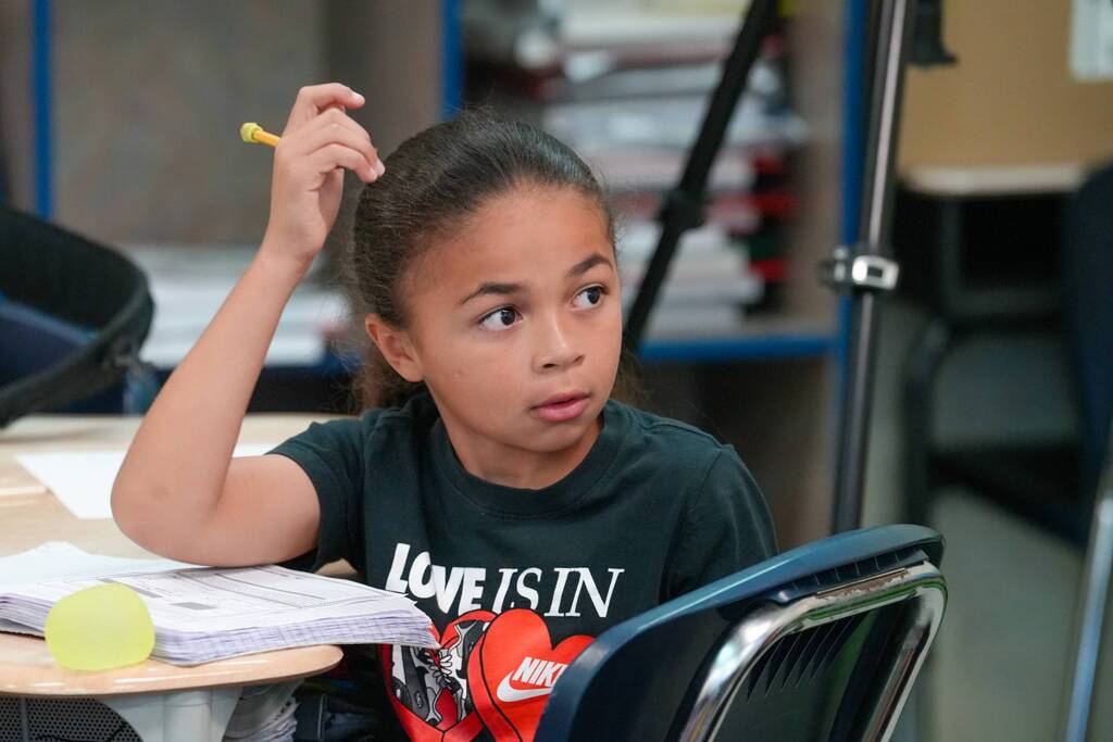 A young girl sits at a desk with a pencil in hand, looking thoughtful, in a classroom setting.