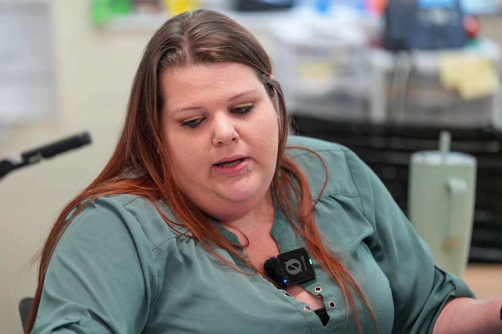 A woman with brown hair sits at a desk, looking down, wearing a green blouse. A microphone and a cup are behind her.