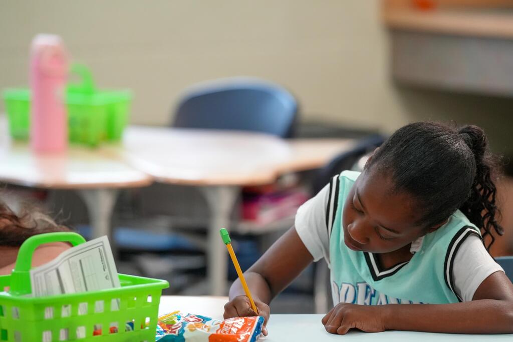 A girl in a classroom with a pencil and paper, surrounded by tables and chairs.