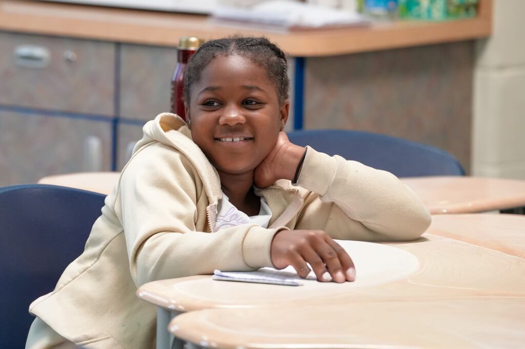 A person in a light beige hoodie sits at a desk in a classroom, smiling and leaning on the table.