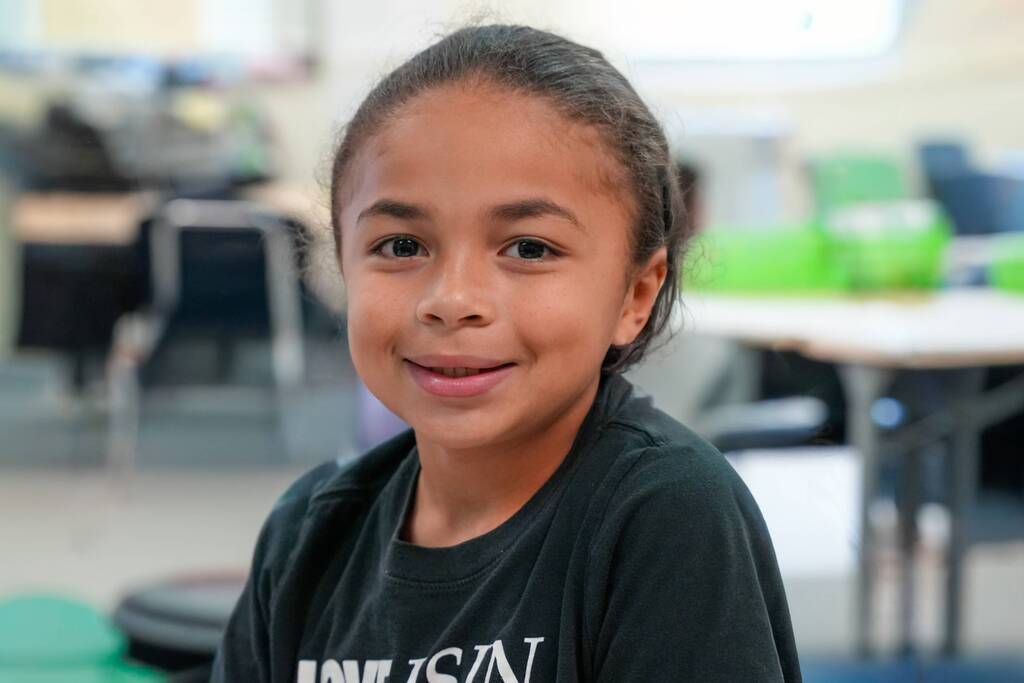 A girl in a black shirt smiles while sitting in a classroom with tables and chairs.