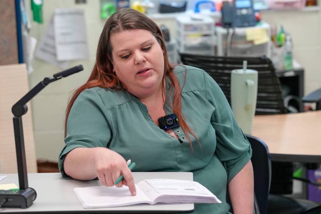 A person in green sits at a desk, reading an open book, with a microphone and other objects in the background.