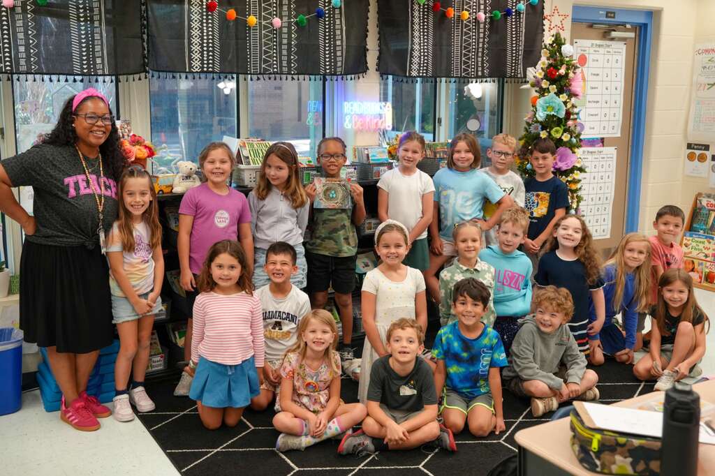 A group of children wearing various casual clothes posing for a photo with an adult in a classroom.