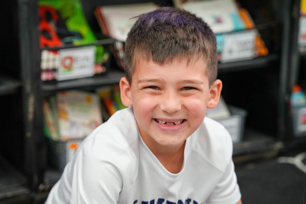 A boy with short hair is smiling in a room with shelves. He is wearing a white shirt.