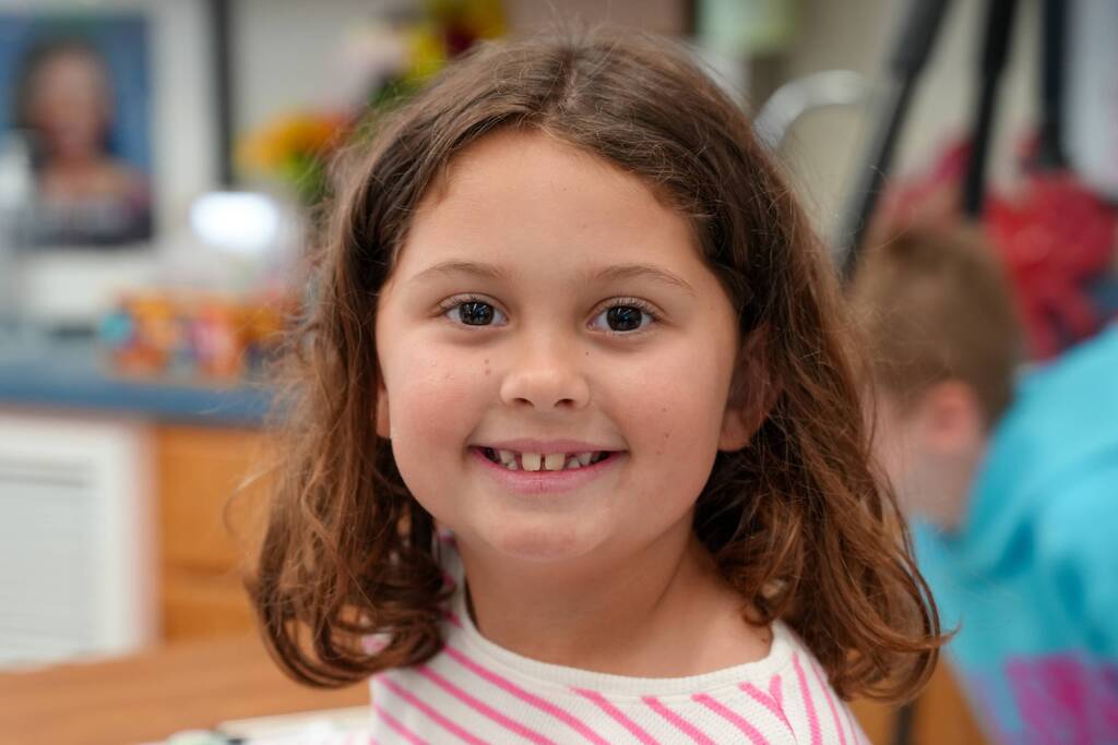A girl with brown wavy hair smiles at the camera. She wears a pink and white striped shirt. A blurred background shows part of another person.