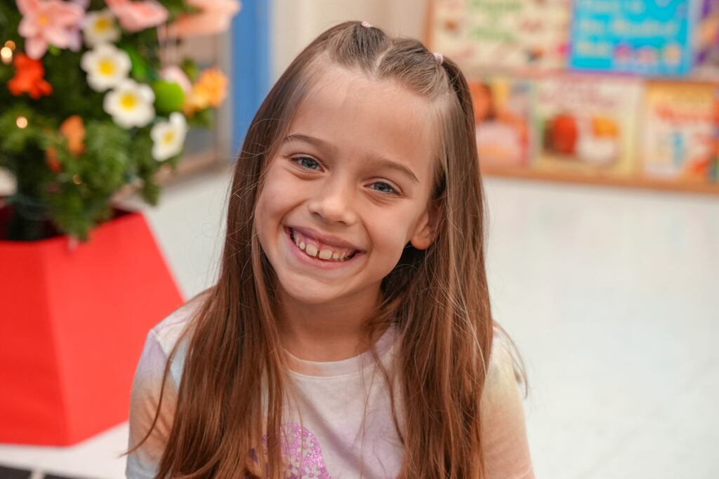 A smiling girl with long hair sits next to a plant in a red planter.