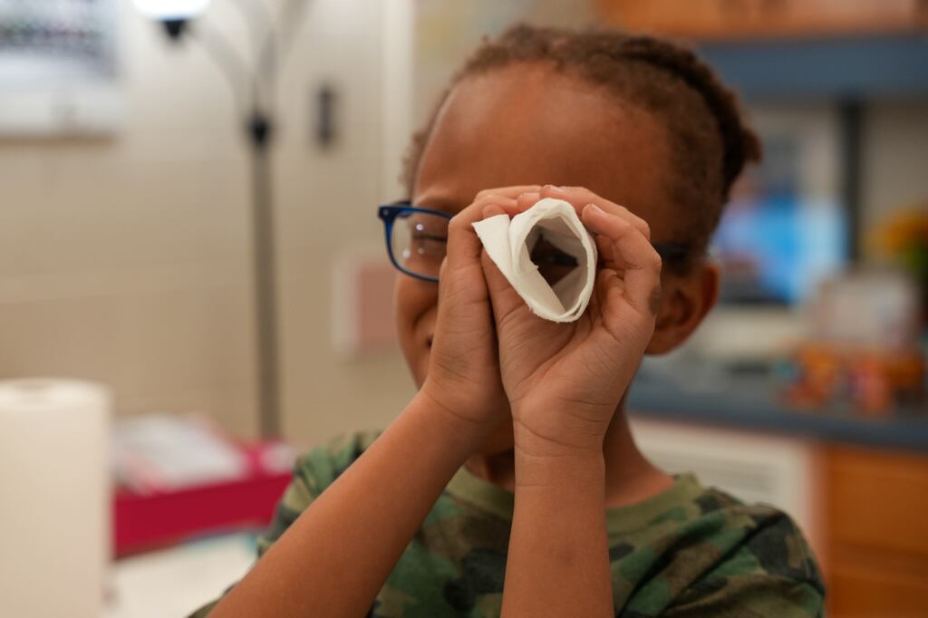 A child with glasses holds a tissue over their face. Behind, a kitchen with a countertop and a plant.